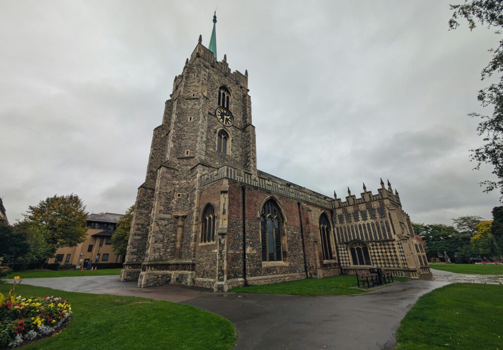 A low-angle view of Chelmsford Cathedral with its clock tower and stained glass windows under a cloudy sky.